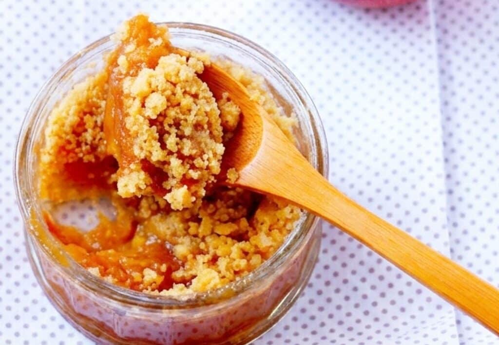 Crumble aux pommes maison dans un petit pot en verre avec une cuillère en bois et une pomme fraîche sur fond clair